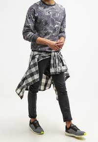 Man in een paarse gemarmerde sweater, zwarte slim fit broek, grijze sneakers met neongele zolen, en een zwart-wit geblokt shirt om zijn taille knopen.