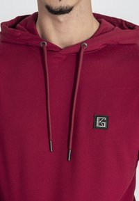 Bordový mikina s kapucňou a šnúrkami, s čiernymi detailmi, so malou vyšitou nášivkou na prednej strane. Hladká textura, ležérny dizajn.