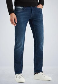 Jeans de mezclilla azul oscuro con un corte ajustado, textura desgastada y diseño clásico de cinco bolsillos. Combinados con zapatillas blancas para un contraste.