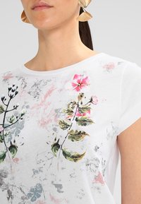 Bílý tričko s květinovým potiskem v růžové a zelené barvě s jemně šedými akcenty. Krátké rukávy a kulatý výstřih. Jemná bavlněná tkanina.