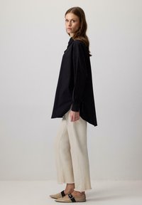 Donna in piedi di lato, con una camicia nera a maniche lunghe e larghi, pantaloni crema a vita alta con orli sfrangiati e ballerine beige con strappi neri.