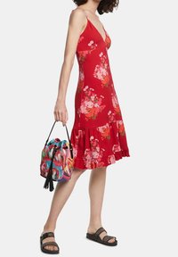 Donna che indossa un vestito senza maniche rosso a fiori e sandali neri, mentre tiene in mano una borsa a secchiello colorata con un motivo astratto e pompon.
