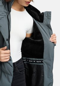 Šviesiai pilkas paminkštintas parka su juodu pūkuotu vidumi, ziperių uždarymu ir „ragwear“ ženklinimu pamušale. Matomas baltas prigludęs marškinys viduje.
