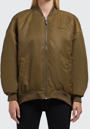 Chaqueta bomber verde oliva con cremallera frontal, puños y dobladillo acanalados y bolsillos laterales, llevada por una persona con pantalones negros.