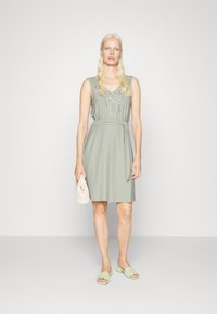 Vestido verde sem mangas com bordados de renda no decote, cintura cingida e um design simples. Combinado com sandálias verde-claro e uma clutch.