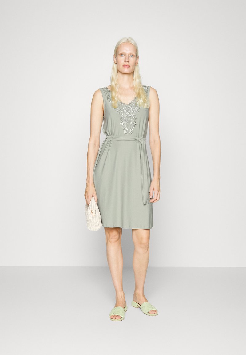 Vestido verde sem mangas com bordados de renda no decote, cintura cingida e um design simples. Combinado com sandálias verde-claro e uma clutch.