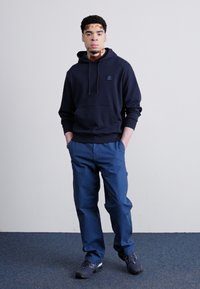 Granatowa bluza z kapturem i sznurkiem, duża kieszeń z przodu oraz logo. Niebieskie spodnie o luźnym kroju. Czarne sneakersy z fioletowymi akcentami.