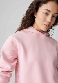 Felpa rosa con cappuccio, realizzata in tessuto morbido. Presenta una vestibilità comoda, polsini a costine e dettagli di cucitura sulle maniche.