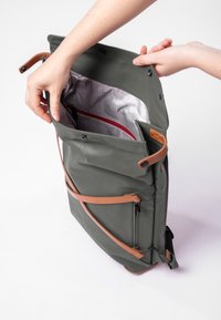 Olivgrüner Rucksack mit Lederakzenten, ausgestattet mit einem Rolltop-Verschluss und einem karierten Innenfutter, enthält eine kleine rote Reißverschluss-Tasche innen.