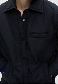 Veste noire à boutons avec col, poche poitrine et mains dans les poches avant sur une personne.