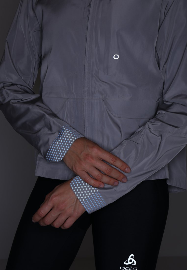 Hellegraue Windjacke mit einem Reißverschluss vorne, die blaue Pünktchenakzente an den Bündchen und ein reflektierendes Logo auf der Brust hat.