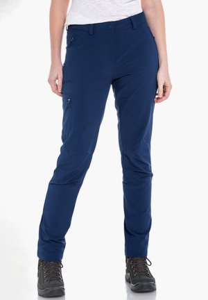 Kobieta ubrana w granatowe spodnie outdoor z kieszeniami na zamki i czarne buty trekkingowe, stojąca na białym tle.