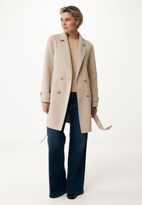 Abrigo de mezcla de lana beige de doble botonadura con botones redondos, solapas con muesca y lazos laterales, combinado con jeans de mezclilla oscuros de pierna ancha.