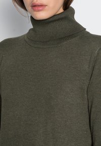 Pull à col roulé vert olive avec une texture côtelée, présentant un col haut et une silhouette ajustée. Tissu léger et doux.