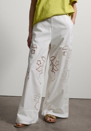 Person trägt weiße Hosen mit weitem Bein und floraler Cutout-Stickerei, ein gelbes Oberteil und gewebte Sandalen, steht vor einem schlichten Hintergrund.