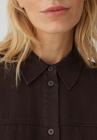 Donna bionda con labbra naturali che indossa una camicia marrone scuro con colletto e bottoni, mostrata dal mento fino alla parte superiore del petto.