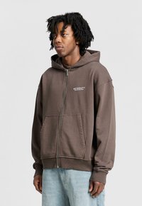 Brown zip-up hoodie featuring a kangaroo pocket, ribbed cuffs, and an embroidered logo on the chest. Soft fabric with a relaxed fit.