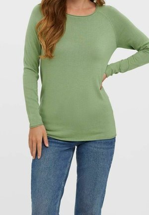 Vrouw draagt een lichtgroen shirt met lange mouwen en een blauwe spijkerbroek, met de ene hand op haar heup en de andere langs haar zij rustend.