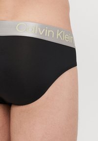 Schwarze Baumwollslips mit einem silbernen elastischen Bund, der das "Calvin Klein"-Logo in hellgrün trägt. Glatte Textur, niedriger Schnitt.