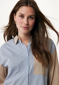 Camicia a righe verticali con un corpo azzurro chiaro e accenti a righe beige. Chiusura con bottoni frontale, collo a contrasto e taschino applicato.