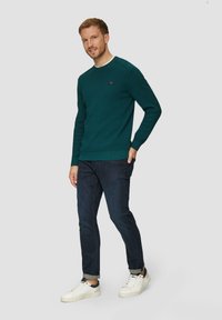 Groene gebreide sweatshirt met een ronde halslijn, geribbelde boorden en een klein logo op de borst, gecombineerd met donkere jeans en witte sneakers.