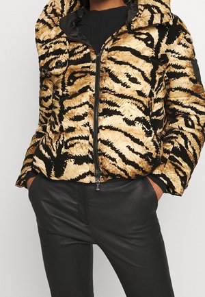 Personne portant une doudoune rayée tigre avec les mains dans les poches d'un pantalon en cuir noir, faisant face à l'avant sur un fond uni.