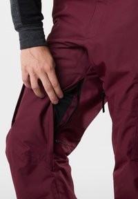 Bordowe spodnie narciarskie z boczną kieszenią na zamek. Wykonane z gładkiego, wodoodpornego materiału. Wewnątrz kieszeni znajduje się teksturowana czarna podszewka.