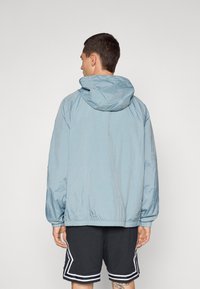 Lyseblå hættejakke med en blank tekstur, elastiske cuffe og hætte med snøre, båret over sorte shorts med hvide detaljer.