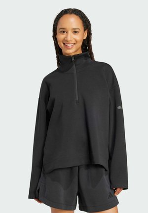 Pull noir avec un col montant et une fermeture zippée quart, présentant des manches amples et des détails côtelés, associé à un short noir.