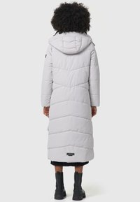 Lange, puffy, grijze winterjas met een capuchon en chevronstiksels. Heeft zwarte accenten en een logo patch dicht bij de zoom. Draagt met zwarte laarzen.
