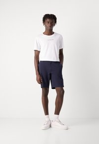 Hvit T-skjorte med "Calvin Klein Jeans"-logo, marineblå shorts og hvite sneakers. Rene linjer, lett stoff, avslappet passform, ankellengde.