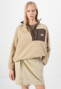 Fleece pullover in beige met een bruine kwartzip en een ritszak op de borst. Gecombineerd met een lichtbeige rok. Gladde textuur.