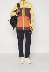 Färgblockerad vattentät jacka i gult, brunt och orange; med dragkedjefickor. Bärs tillsammans med svarta byxor och ljusa skor.