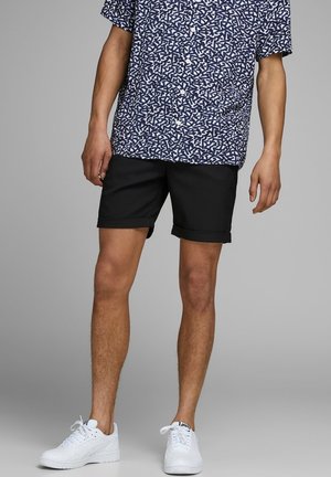 Homme portant une chemise à manches courtes bleu marine à motifs avec des boutons, un short noir à ourlet retroussé et des baskets blanches, debout devant un fond gris.