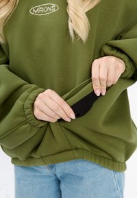 Olivgrüner, übergroßer Sweatshirt mit umgeschlagenen Ärmeln, gesticktem Logo und einer schwarzen Taschenöffnung an der Seite, getragen über hellblauer Jeans.
