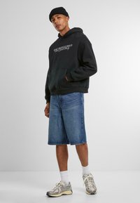 Sort hoodie med frontlomme og tekstdesign, parret med knæ-lange denim shorts og grå løbesko med hvide accenter.