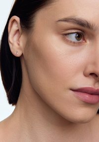 Złoty kolczyk w kształcie słuchawki z małym, łzawym akcentem przypominającym diament. Prosty design na neutralnym odcieniu skóry. Widoczna gładka tekstura.