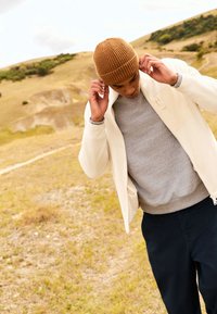 Cremefarbene leichte Jacke mit Reißverschluss, grauer Pullover darunter, marineblaue Hose und braune, gerippte Mütze vor einer grasbewachsenen Landschaft.