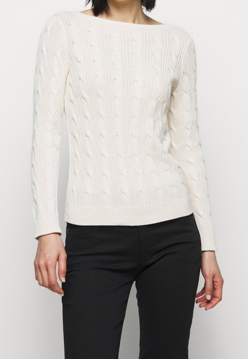 Personne portant un pull en maille torsadée blanc à manches longues et un pantalon noir sur un fond clair uni.