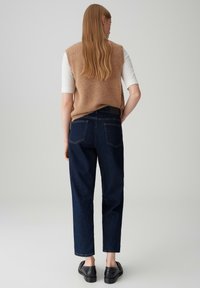 Mouwloos beige gebreide trui over een wit shirt, gecombineerd met donkerblauwe high-waisted jeans met achterzakken en zichtbare stiksels.