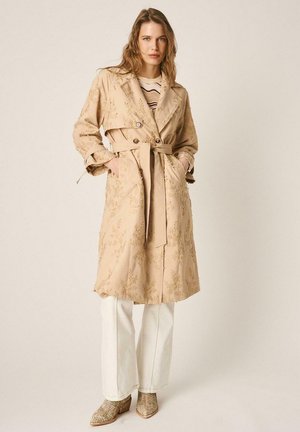 Mujer con un abrigo trench beige bordado sobre una camiseta de rayas, pantalones blancos de pierna ancha y botines estampados, de pie con las manos en los bolsillos.