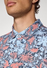 Short-sleeve polo shirt in blue with a floral pattern in red and black. Features a classic collar and buttons, with an embroidered logo accent.