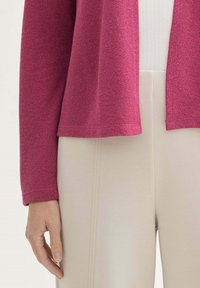 Fuchsia stickad cardigan med en avslappnad passform, öppen framsida och långärmar, kombinerad med beigea tighta byxor med vertikala sömdetaljer.