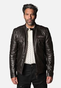 Veste en cuir marron avec des accents matelassés sur les épaules, deux poches frontales zippées et un logo discret. Portée sur une chemise blanche texturée.