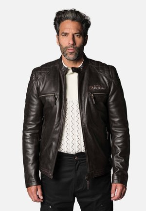 Veste en cuir marron avec des accents matelassés sur les épaules, deux poches frontales zippées et un logo discret. Portée sur une chemise blanche texturée.