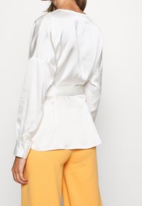 Vit satinklänning med långa ärmar, midjeband och mjuka draperingar, med rund halsringning och diskret lyster. Matchas med gula byxor.