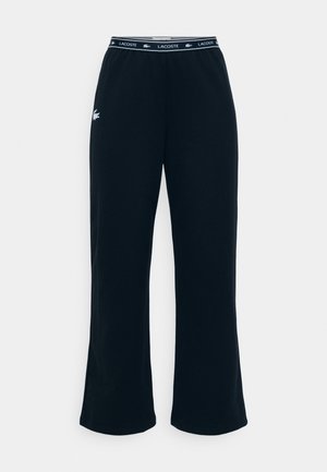 Pantalones de chándal negros con un diseño de pierna acampanada, fabricados en tela suave. Presentan un logo blanco de Lacoste en la cadera izquierda y una cinturilla con marca.