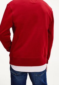 Red sweatshirt made of soft cotton, featuring a crew neck, long sleeves, and a ribbed hem. Worn over a white layered shirt and blue jeans.
