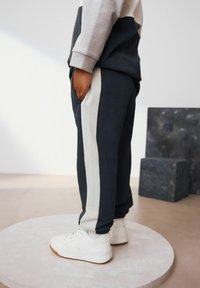 Pantaloni della tuta grigio scuro con strisce bianche ai lati, abbinati a sneaker bianche. Il tessuto sembra morbido e casual, con una vita elasticizzata.