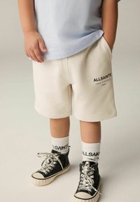 Pantaloni corti in cotone beige chiaro con tasche laterali e logo "ALLSAINTS". Abbinati a sneaker alte nere e calze bianche a coste con logo.
